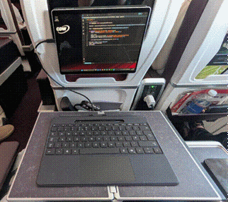 Title: Surface Pro wedged into an airplane seat-back with the keyboard detached - Description: The Surface Pro screen wedged into the seat-back recess showing code, keyboard detached and resting on the tray in front, on board a flight.